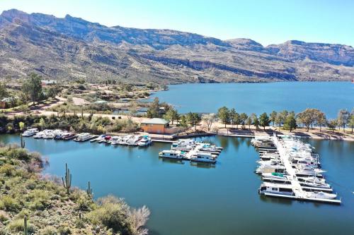 Aerial view of a marina with boats on a calm lake, surrounded by mountains and greenery under a clear blue sky.