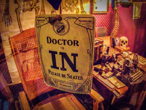 A vintage "Doctor is IN" sign hangs in a cluttered room with a skeleton, books, and old medical tools.