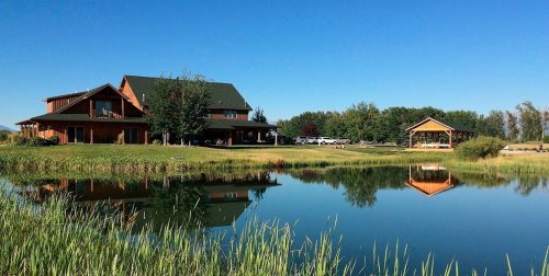 A large wooden house and gazebo reflect in a calm pond, surrounded by green grass and trees under a clear blue sky.