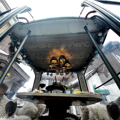 A cozy dining setup inside a glass structure, featuring a table with food, wine, and a chandelier above.