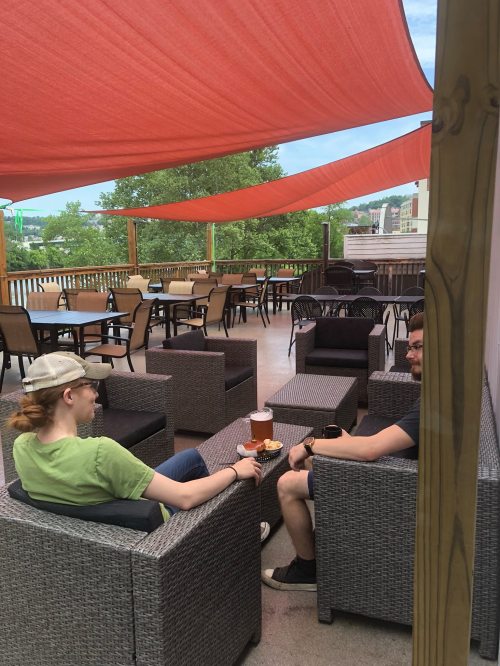Two people sit on a patio under a red canopy, enjoying drinks and snacks in a relaxed outdoor setting.