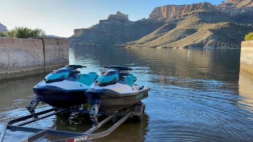Two jet skis on a trailer by a calm lake, surrounded by mountains and clear blue skies.
