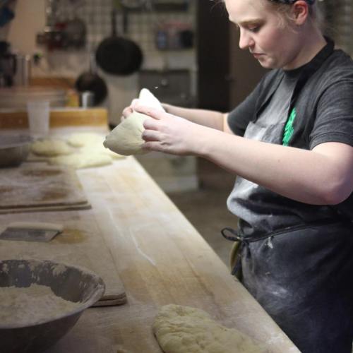 A baker shapes dough on a wooden countertop, surrounded by flour and baking tools in a cozy kitchen setting.