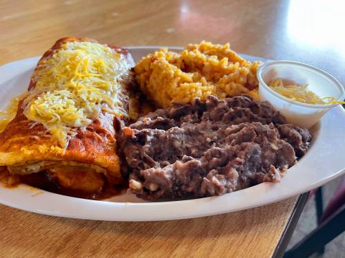 A plate of enchiladas topped with cheese, served with rice and refried beans on a wooden table.