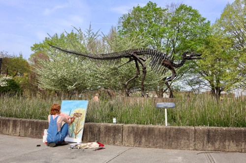A person paints in front of a dinosaur skeleton surrounded by greenery on a sunny day.