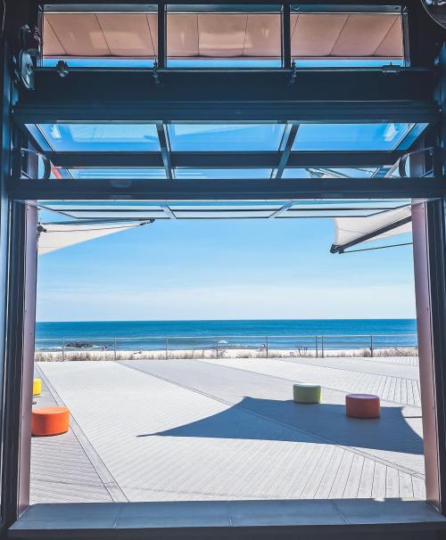 A view of the ocean through large glass doors, with colorful seating on a wooden deck under a clear blue sky.