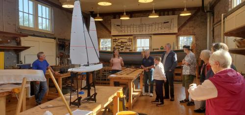A group of people gathers in a workshop with model boats and wooden materials, discussing boat building.