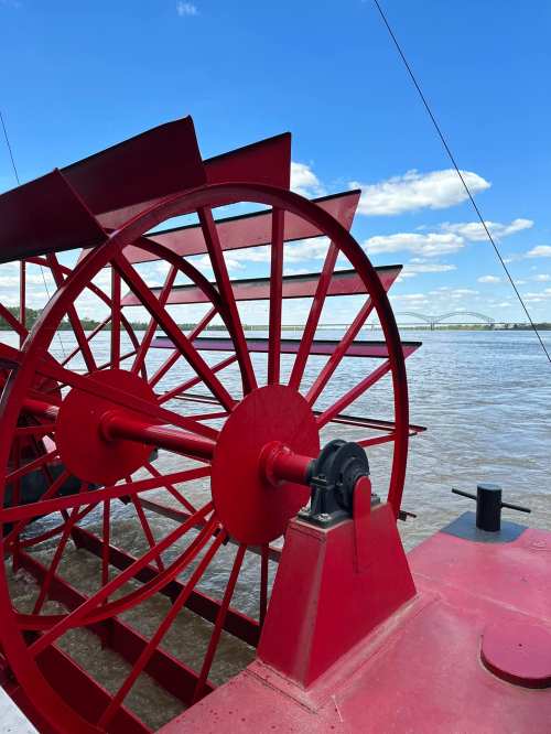 Close-up of a red paddle wheel on a riverboat, with a blue sky and water in the background.