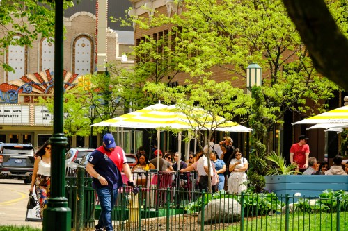 A lively outdoor scene with people dining under umbrellas, surrounded by greenery and a theater marquee in the background.
