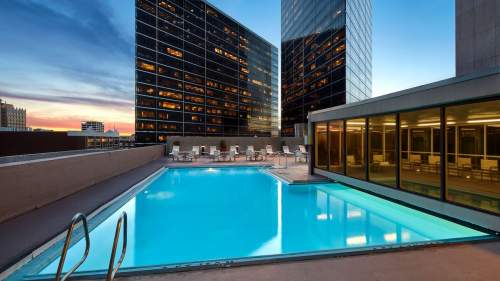 Rooftop pool surrounded by modern buildings at sunset, with lounge chairs and a clear blue sky.