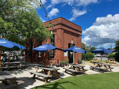 A brick building with blue umbrellas over picnic tables, surrounded by greenery and a bright blue sky with clouds.