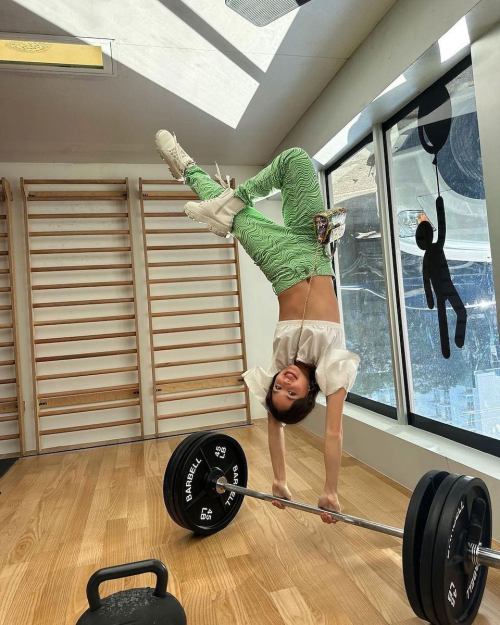 A person performs a handstand on a barbell in a gym, wearing green striped pants and a white top.