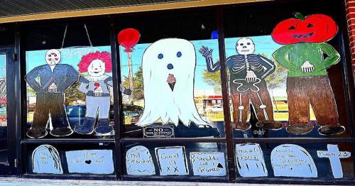 Window display featuring Halloween-themed characters: a ghost, skeleton, clown, and pumpkin, with playful decorations.