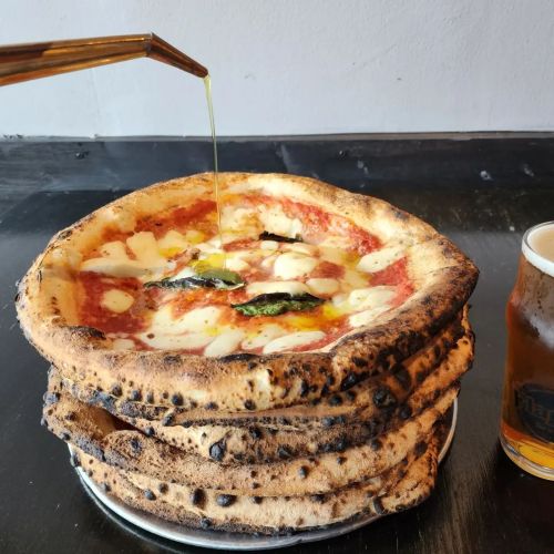 A stack of pizzas with melted cheese and basil, drizzled with oil, next to a glass of beer on a black table.