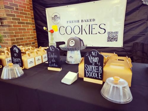 A table displaying fresh baked cookies with labeled containers, a hat, and a banner in the background.