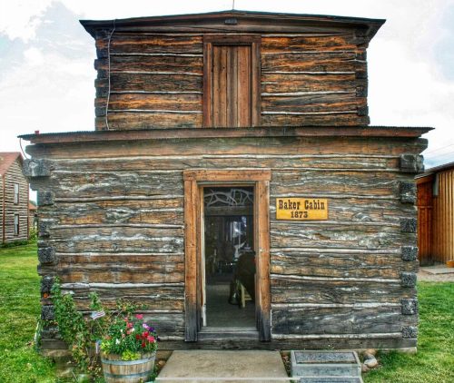 A rustic wooden cabin with a sign reading "Bakery Cabin 1873," surrounded by greenery and flowers.