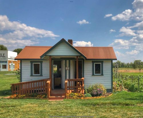 A small blue house with a brown metal roof, surrounded by greenery and flowers under a partly cloudy sky.