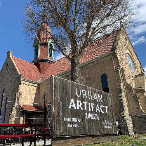 A historic building with a tall steeple, featuring a sign for "Urban Artifact Taproom" and outdoor seating.