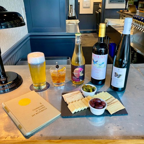 A selection of drinks and snacks on a table, including beer, wine, a cocktail, olives, and crackers.