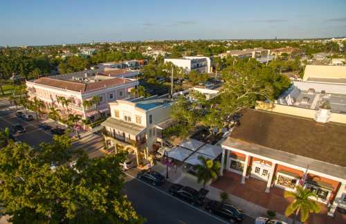 Aerial view of a vibrant town with shops, palm trees, and parked cars under a clear blue sky.