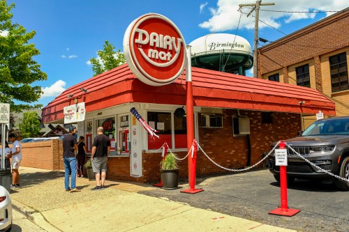 A retro-style ice cream shop with a red roof and sign, featuring customers in line on a sunny day.