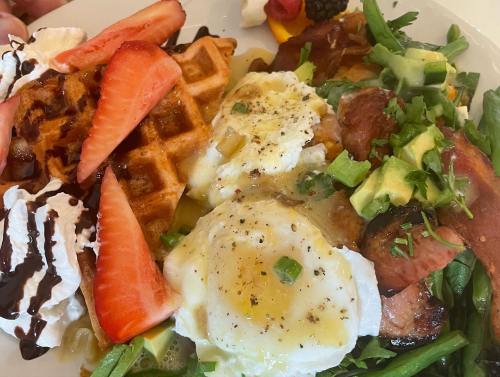 A plate of waffles topped with strawberries, whipped cream, and a savory dish with poached eggs, bacon, and avocado.