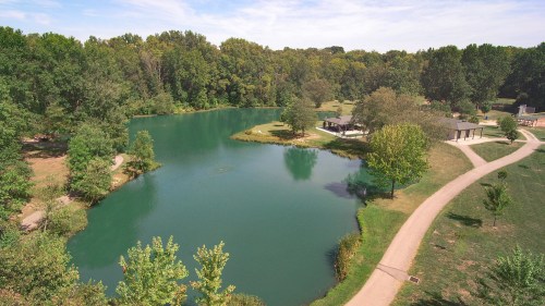 A serene park scene featuring a calm lake surrounded by trees and walking paths on a sunny day.