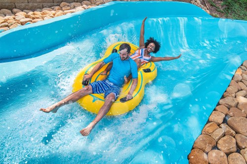 A man and woman joyfully ride a yellow inflatable tube down a water slide, splashing in the blue water.