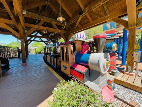 A colorful miniature train sits under a wooden structure, surrounded by greenery and bright sunlight.