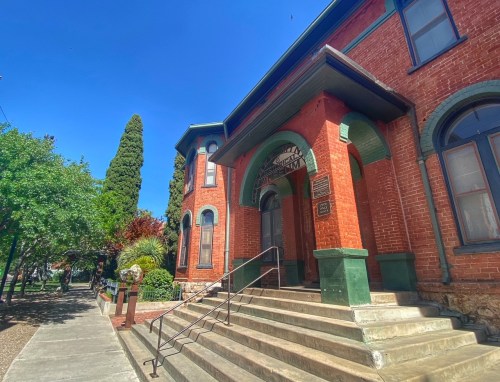 Historic brick building with green accents, steps leading up, surrounded by trees and clear blue sky.