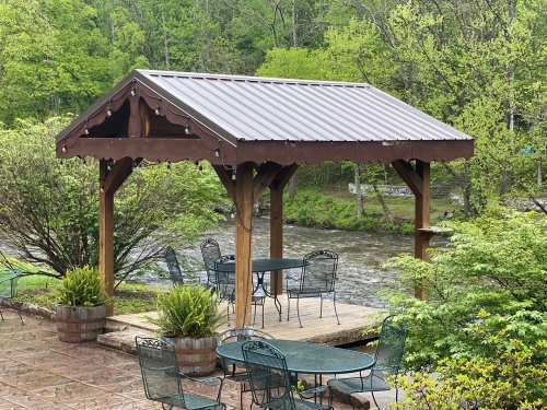 A wooden gazebo with a metal roof beside a river, surrounded by greenery and outdoor seating.