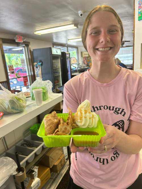 A smiling woman holds a green tray with desserts, including a swirl of soft serve and two pastries.