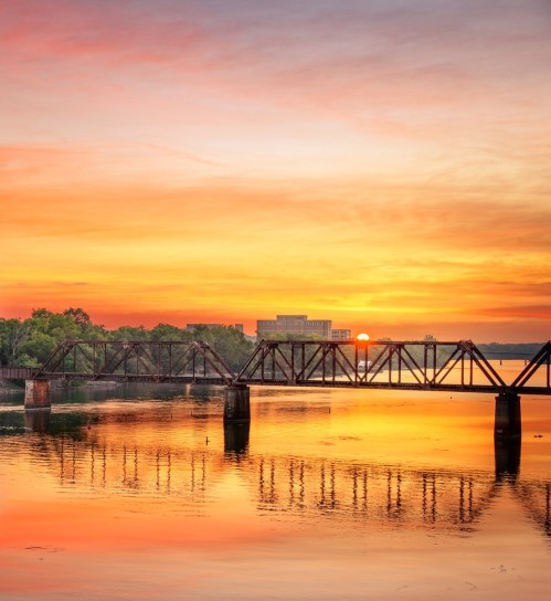 Sunset over a river with a silhouetted bridge and vibrant orange and pink sky reflecting on the water.