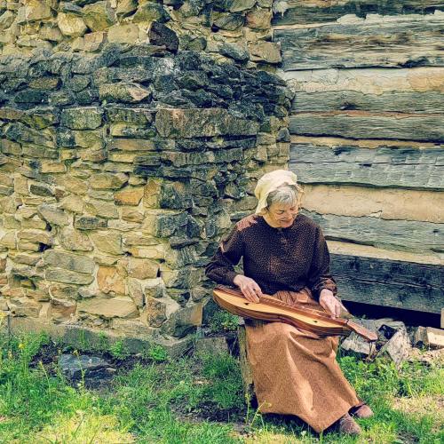 An elderly woman in period clothing plays a dulcimer beside a stone wall and wooden cabin.