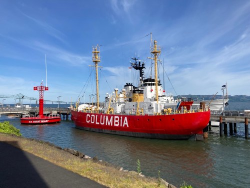 A red and white ship named "Columbia" docked near a red lighthouse with a U.S. Coast Guard sign. Clear blue sky above.