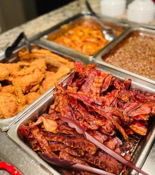 A buffet spread featuring crispy fried chicken, bacon strips, and a sweet dessert in metal trays.