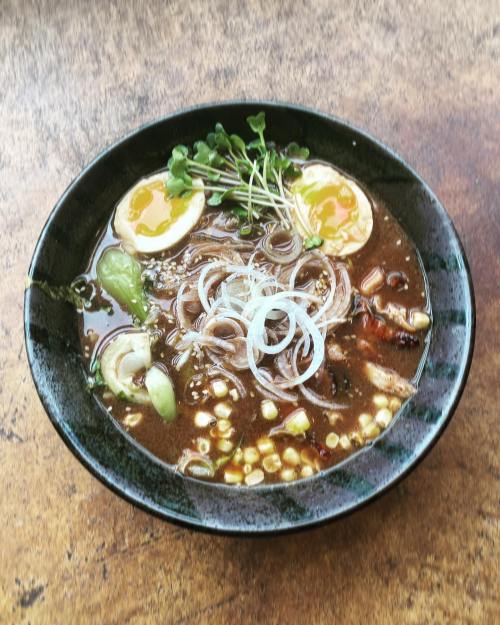 A bowl of ramen with broth, noodles, sliced eggs, green onions, and vegetables, served in a dark bowl on a wooden table.