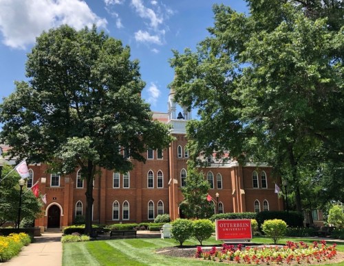A brick university building surrounded by trees and colorful flowers, with a sign for Otterbein University in front.