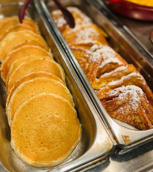 A tray of golden pancakes next to a tray of pastry drizzled with powdered sugar.