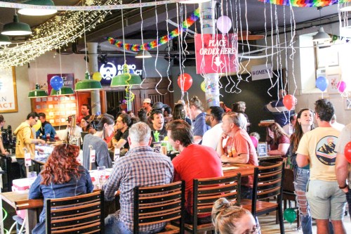 A lively bar scene with people socializing, colorful decorations, balloons, and a festive atmosphere.
