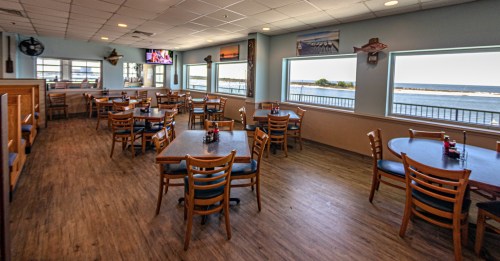 A spacious restaurant interior with wooden tables and chairs, large windows overlooking a waterfront view.