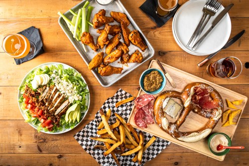 A spread of food including wings, a salad, pretzel, charcuterie, fries, and drinks on a wooden table.