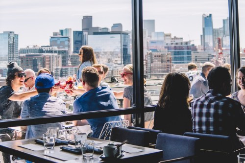 A lively rooftop gathering with people enjoying drinks and city views on a sunny day.