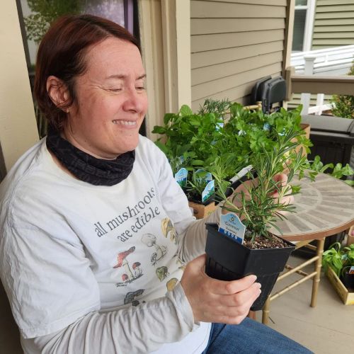 A person smiles while holding a potted rosemary plant on a porch, surrounded by greenery.