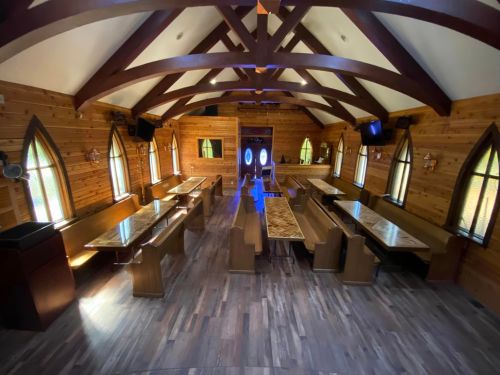 Interior of a wooden chapel with arched beams, long tables, and stained glass windows, featuring a spacious layout.