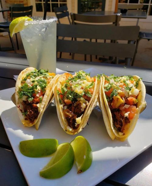 Three tacos filled with meat, topped with cilantro, tomatoes, and cheese, served with a drink and lime wedges.