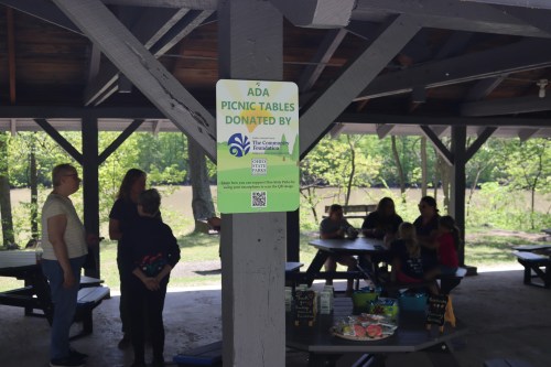A sign about ADA picnic tables hangs in a pavilion, with people gathered around picnic tables in the background.