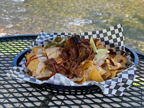 A basket of nachos topped with pulled beef, cheese sauce, jalapeños, and barbecue sauce, set against a scenic background.