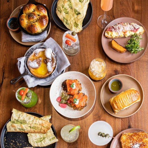 A variety of dishes on wooden table, including bread, pastries, salads, and drinks, arranged in colorful plates.
