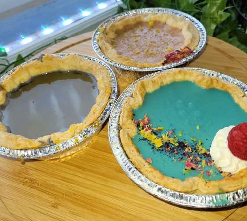 Three colorful mini pies in aluminum crusts: one gray, one teal with a raspberry, and one light purple, on a wooden surface.